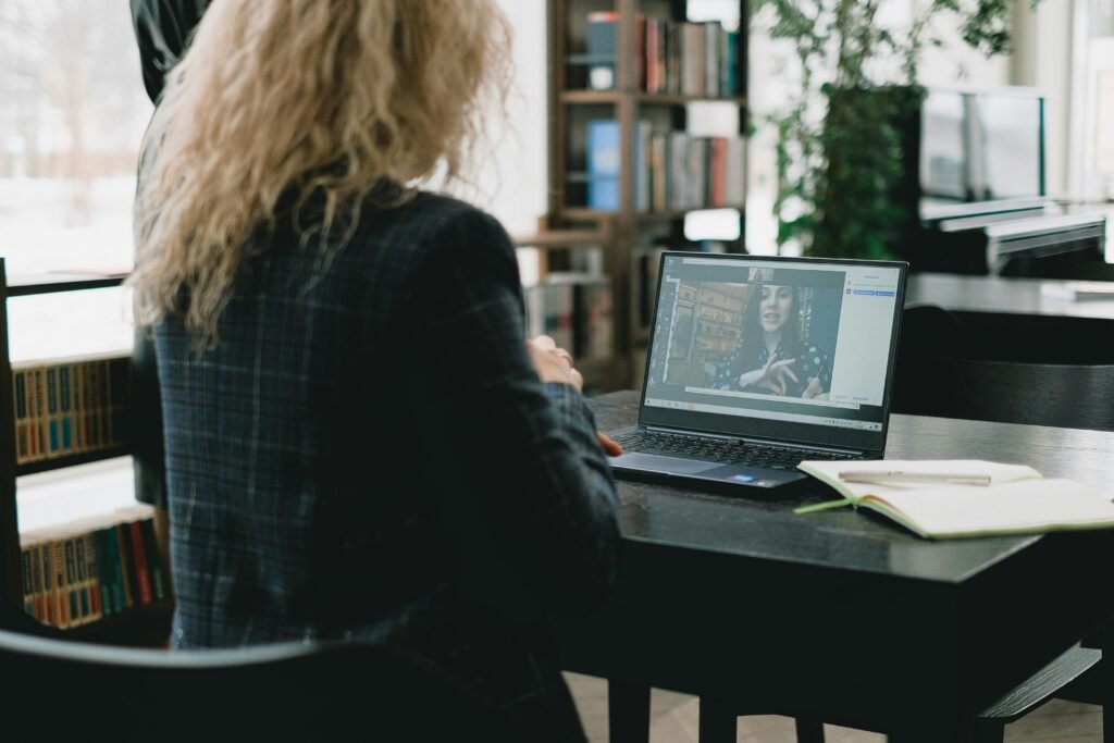A woman sits at a laptop in a stylish library setting, engaging in a video call.